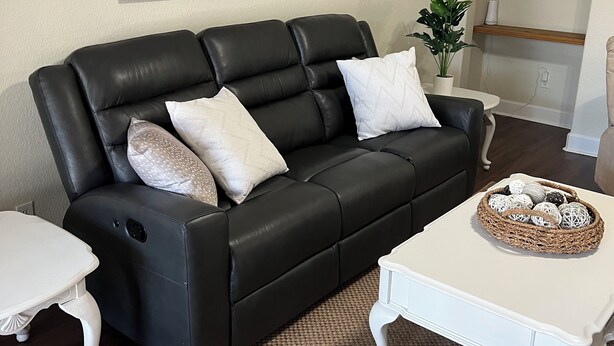 A living room with a black leather sofa, a white coffee table, a woven rug, and a painting of a barn and truck.