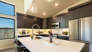 A modern kitchen with dark cabinetry, stainless steel appliances, a large island, and a view through the window. - Free Ski Shuttle- Minutes to Ski and Lake, Community Clubhouse-Pool & Hot Tub (Heber City)