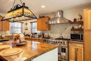 A kitchen with wooden cabinets, a stainless steel range hood, a microwave, and a large island with a stained glass light fixture.