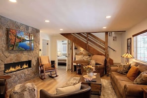 A cozy living room with a stone fireplace, a comfortable sofa, a wooden rocking chair, and a staircase leading upstairs. - Foxy Farm Ranch, guest house, minigolf, hot tub (Big Bear Lake)