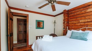 A bedroom with a wooden log wall, a ceiling fan, a bed with white bedding, a blue pillow, and a stack of white towels.