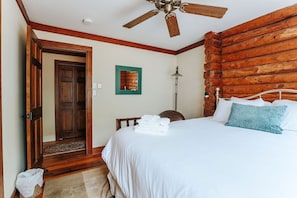 A bedroom with a wooden log wall, a ceiling fan, a bed with white bedding, a blue pillow, and a stack of white towels.