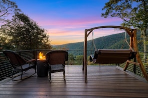 Eine Holzterrasse mit Schaukelstuhl und Schaukel, einem Feuerkorb und Blick auf einen bewaldeten Gebirgszug.