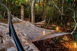 A hammock set up between trees in a wooded area with stone walls and a stone bench.
