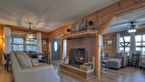 A cozy living room with a fireplace, wooden walls, and a dining area. - Moon Tower Cabin - Beautiful Water Views, Firepit + More! (Blairsville)