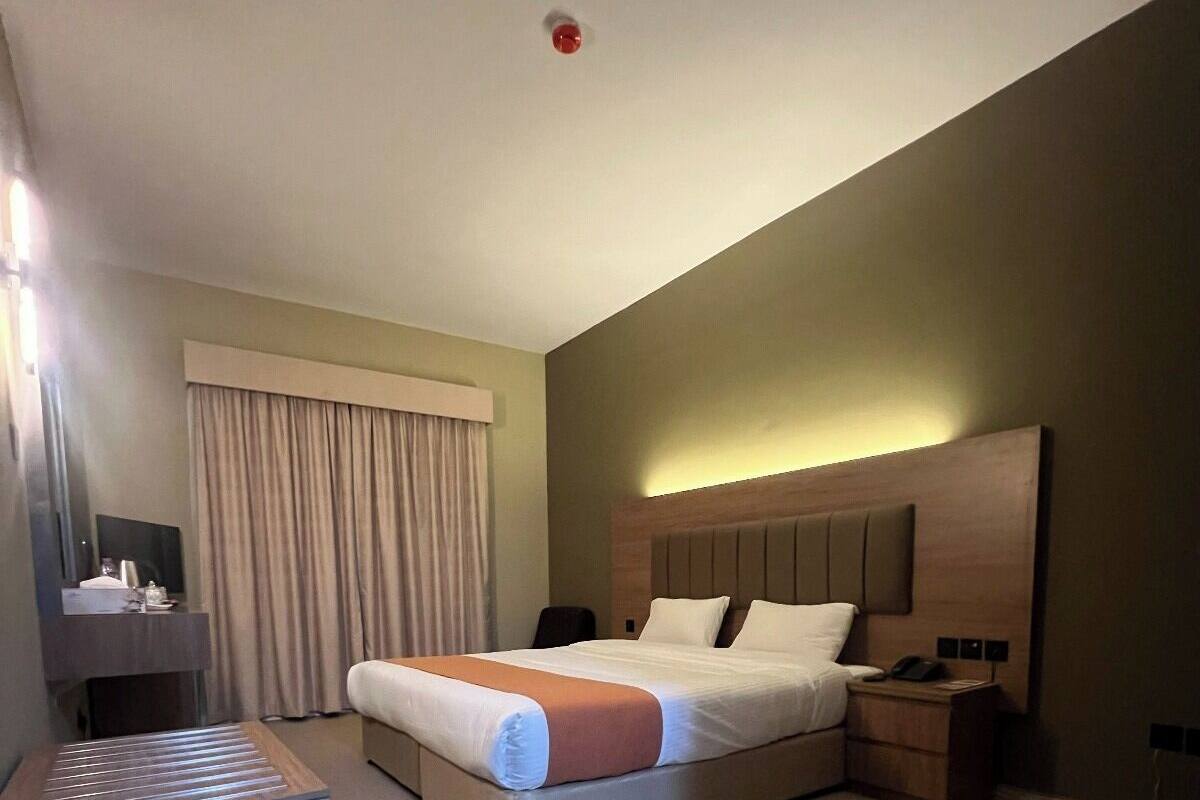 A hotel room with a large bed, a bedside table, a nightstand, a desk with a chair, and a window with curtains.