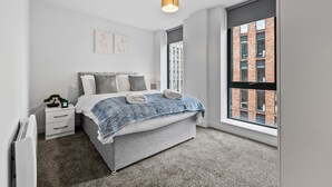A modern bedroom with a large window, a bed with a blue patterned blanket, a bedside table with a plant, and a radiator. - VICHY - Baltic Triangle Modern Apartment (Liverpool)