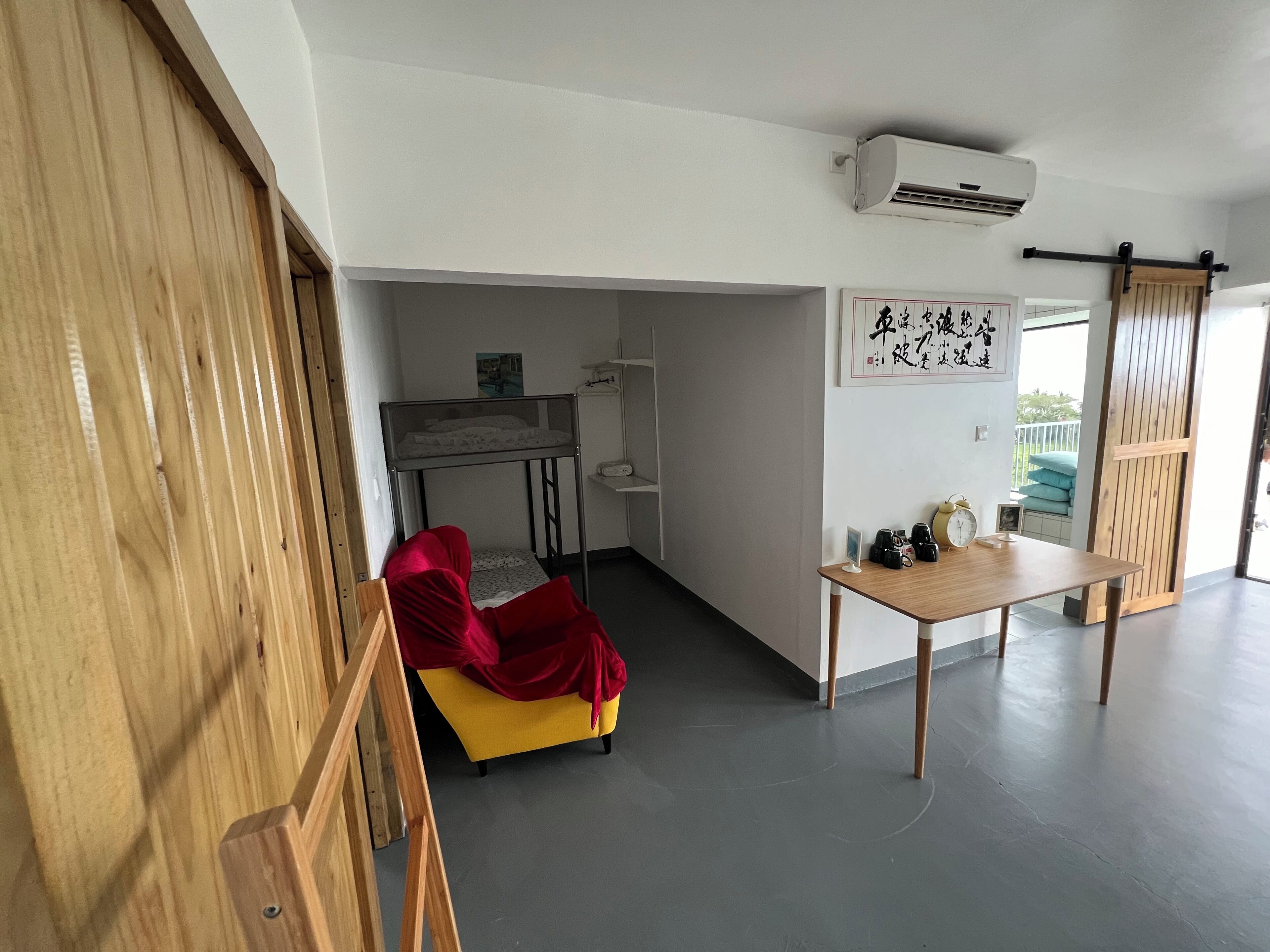 Una habitación con sofá amarillo, manta roja, mesa de madera con objetos, aire acondicionado blanco y puerta corredera.
