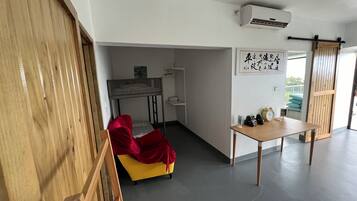 Una habitación con sofá amarillo, manta roja, mesa de madera con objetos, aire acondicionado blanco y puerta corredera.
