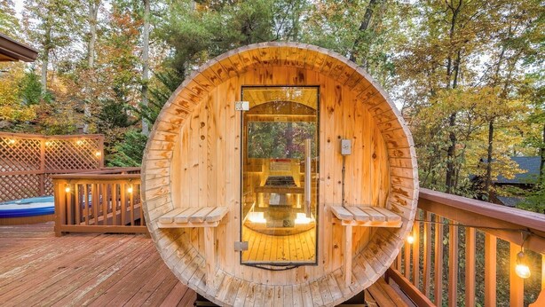 Une structure de sauna en bois, dotée de bancs et d’une porte en verre, entourée d’une terrasse en bois et d’arbres.