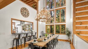 Une salle à manger avec une longue table en bois dressée pour un repas, entourée de chaises. Une grande horloge et un lustre sont visibles sur les murs.