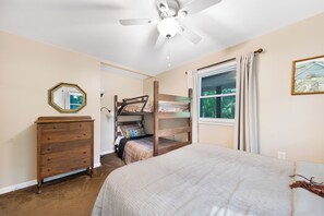 Un dormitorio con una cama litera, un aparador, un ventilador de techo, una ventana con cortinas y un cuadro en la pared.
