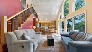 Un amplio espacio de sala con un sofá grande de color gris, un sofá azul y un tapete estampado. Una escalera con barandillas de madera conduce a un nivel superior.