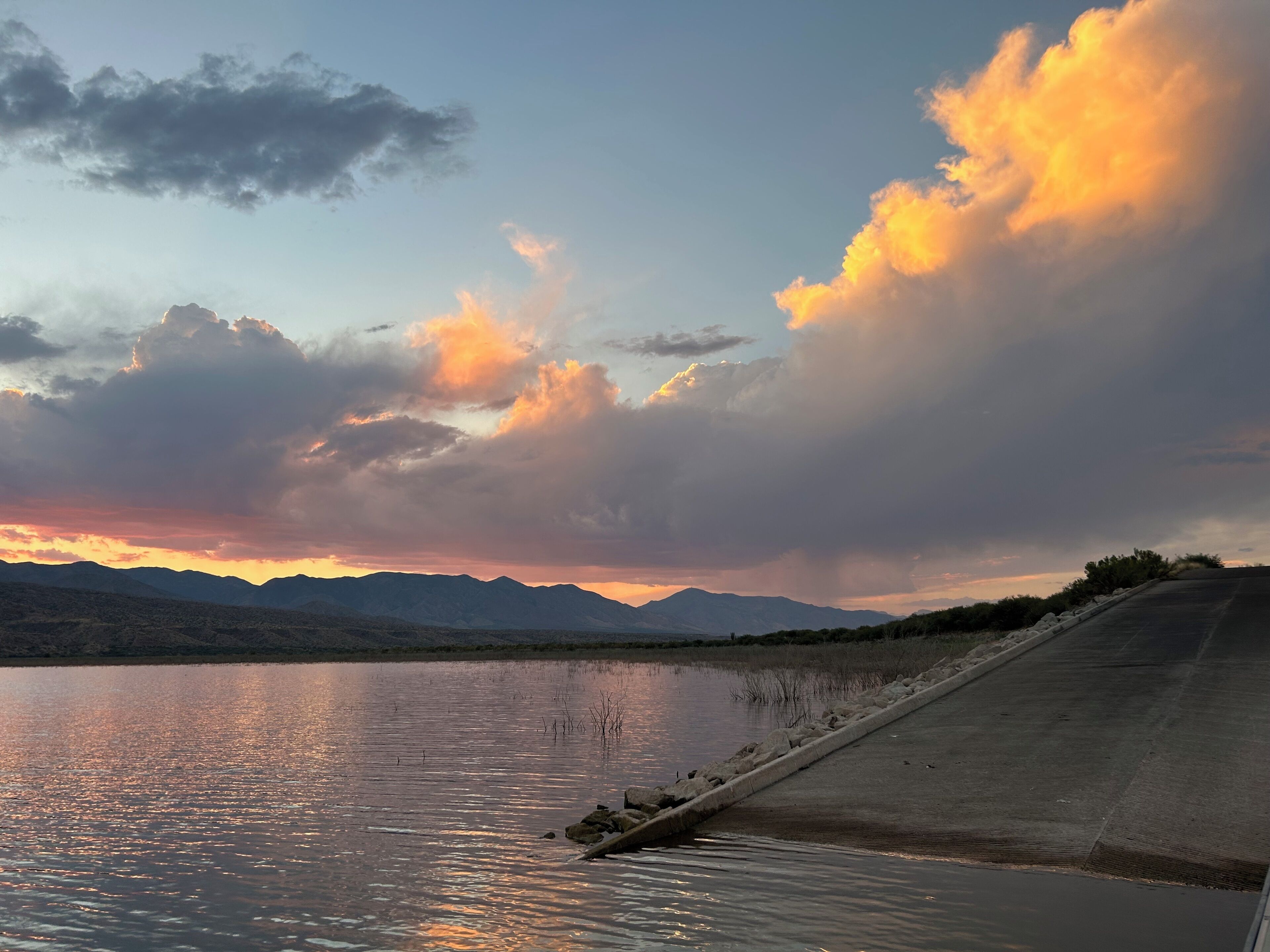 Un lago tranquilo con montañas al fondo y un muro de hormigón a la derecha.