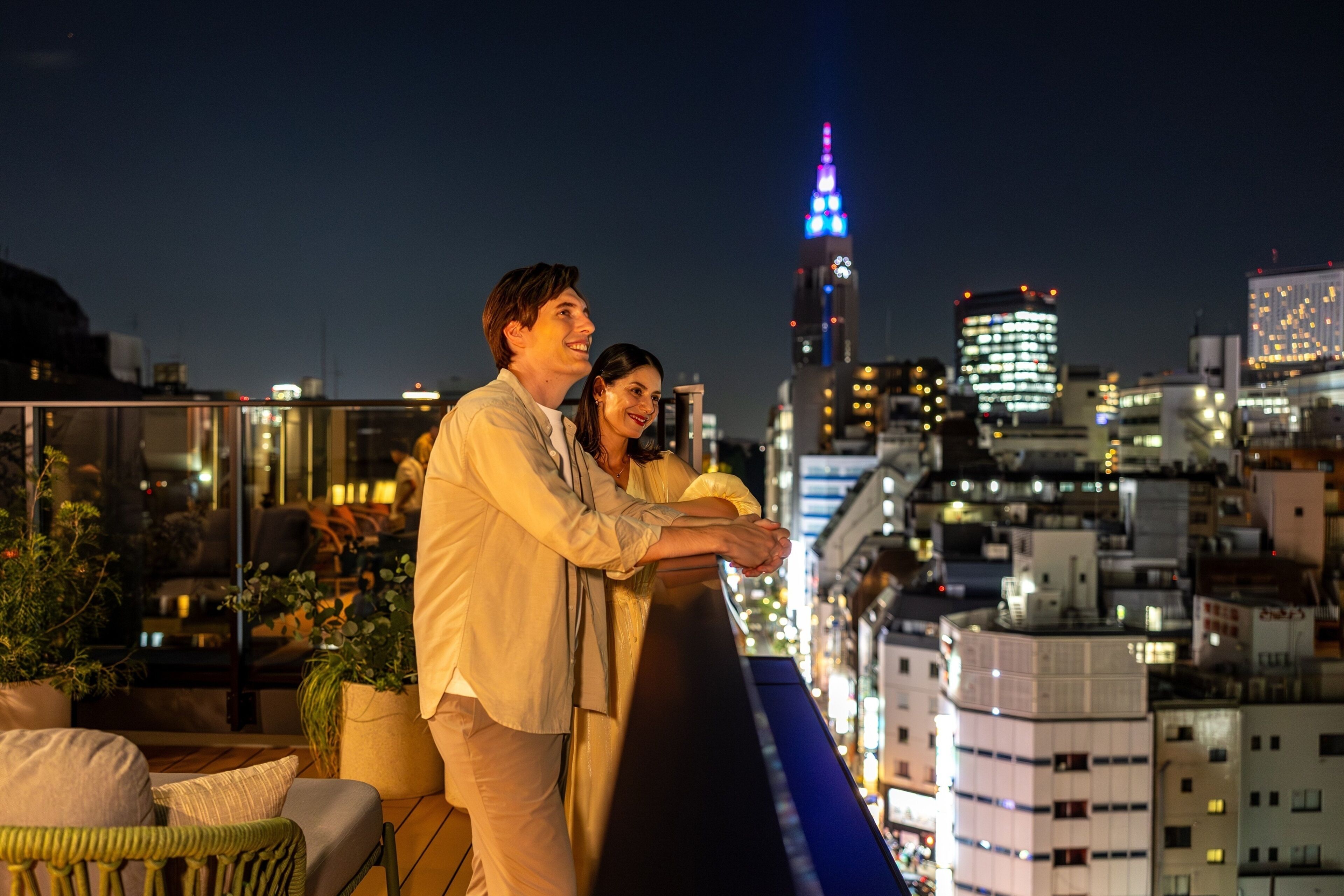 Una pareja en un balcón con vistas a un paisaje urbano, incluyendo un edificio destacado con ventanas iluminadas.