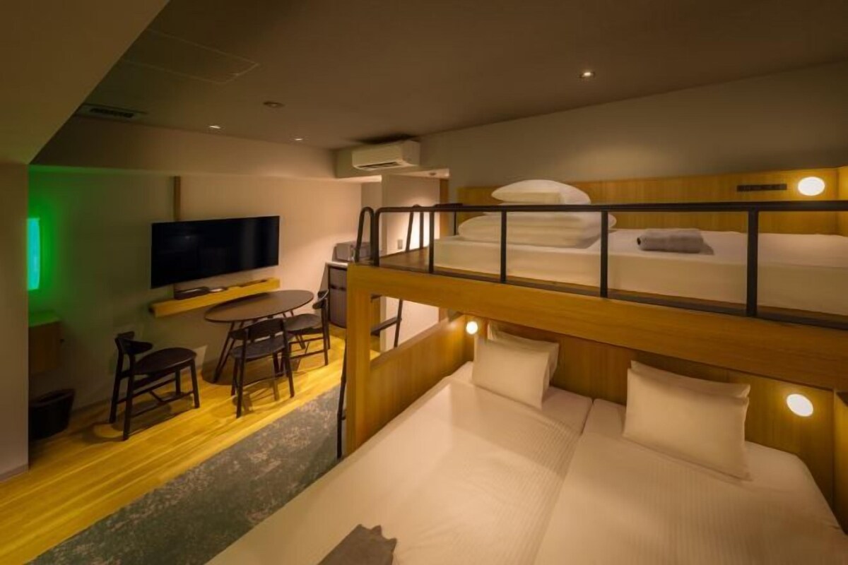 A compact hotel room with a bunk bed, a desk with a chair, a television, and a small seating area with chairs.
