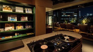 A modern lounge area with a DJ setup, a bookshelf with various items, and a city view. - &Here SHINJUKU (Tokyo)