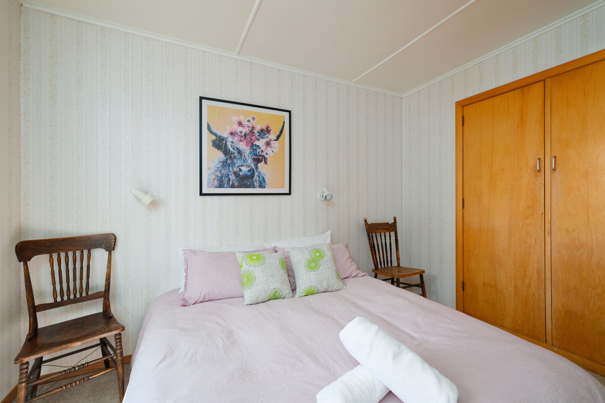 Une chambre à coucher avec un lit, deux chaises en bois, une armoire en bois et un tableau encadré accroché au mur.