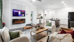 A modern living room with a fireplace, seating area, and a kitchen in the background. - Regal Hideaway | Pool Near Lackland + Med Ctr Gym! (San Antonio)