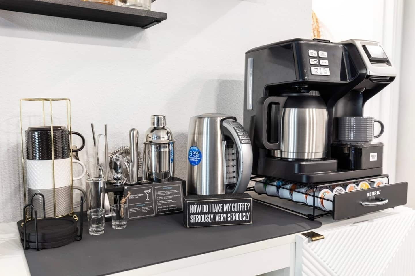 A coffee station with a Keurig coffee maker, a coffee pod holder, a coffee grinder, a milk frother, and a sign asking how to take coffee.