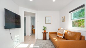 Een moderne woonkamer met een flatscreen televisie aan de muur, een bruine leren bank met een gestreept kussen, een klein rond tafeltje met een potplant en een groot raam met uitzicht op groen.