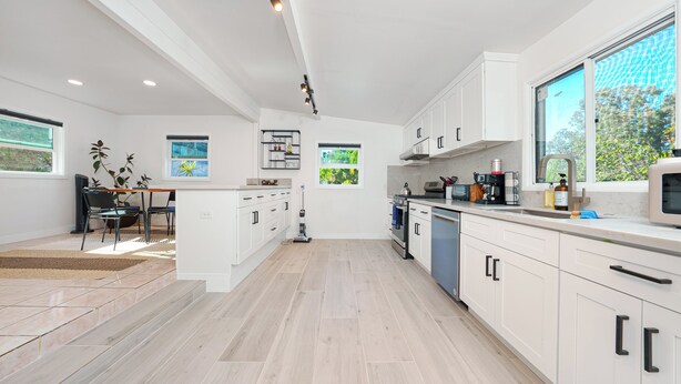 Een moderne keuken met witte kasten, een eethoek met een tafel en stoelen, en een groot raam dat voor natuurlijk licht zorgt.