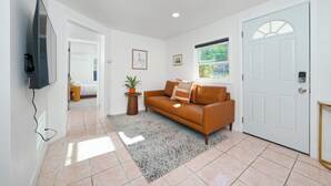Een moderne woonkamer met een bank, een aan de muur bevestigde televisie, een deur en een raam met uitzicht op groen.