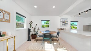 Een moderne keuken met witte kasten, een ronde eettafel en twee ramen met uitzicht op groen.