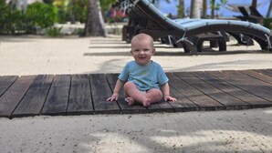 A baby sitting on a wooden deck with a beach and lounge chairs in the background.