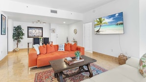 A spacious living room with a large orange sofa, a coffee table, and a tropical beach painting on the wall. - Waterfront Downtown West Palm Beach with  Luxury Amenities (West Palm Beach)