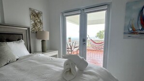 Uma cama bem arrumada com uma toalha enrolada, um criado-mudo com abajur, um quadro na parede e uma porta de vidro deslizante que dá para área externa com uma cadeira vermelha e uma rede.