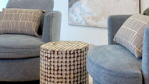 Una stanza con un grande dipinto raffigurante una spiaggia, due divani grigi con cuscini decorati e un tavolino intrecciato.