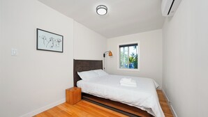 Un dormitorio con cama, mesita de noche, cuadro enmarco, ventana y unidad de aire acondicionado.