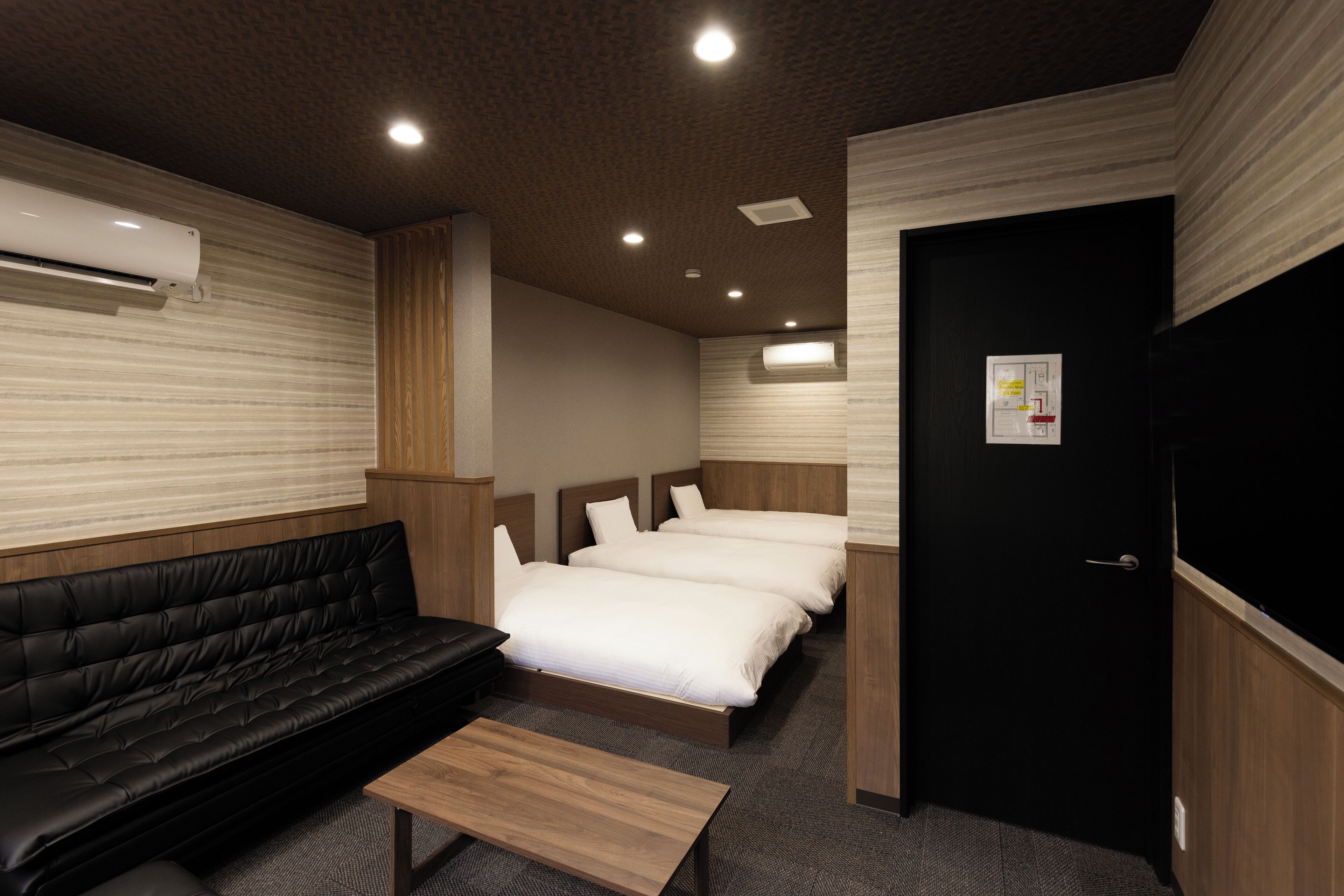 A hotel room with a black leather sofa, two single beds, a wooden bench, and a black door.