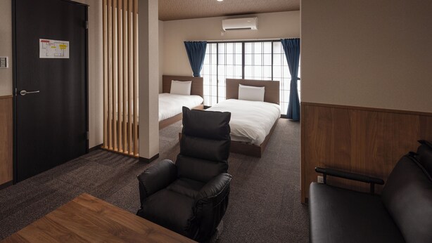 A hotel room with two beds, a wooden desk, a leather armchair, and a wooden partition. - Whitedragon60 (Osaka)