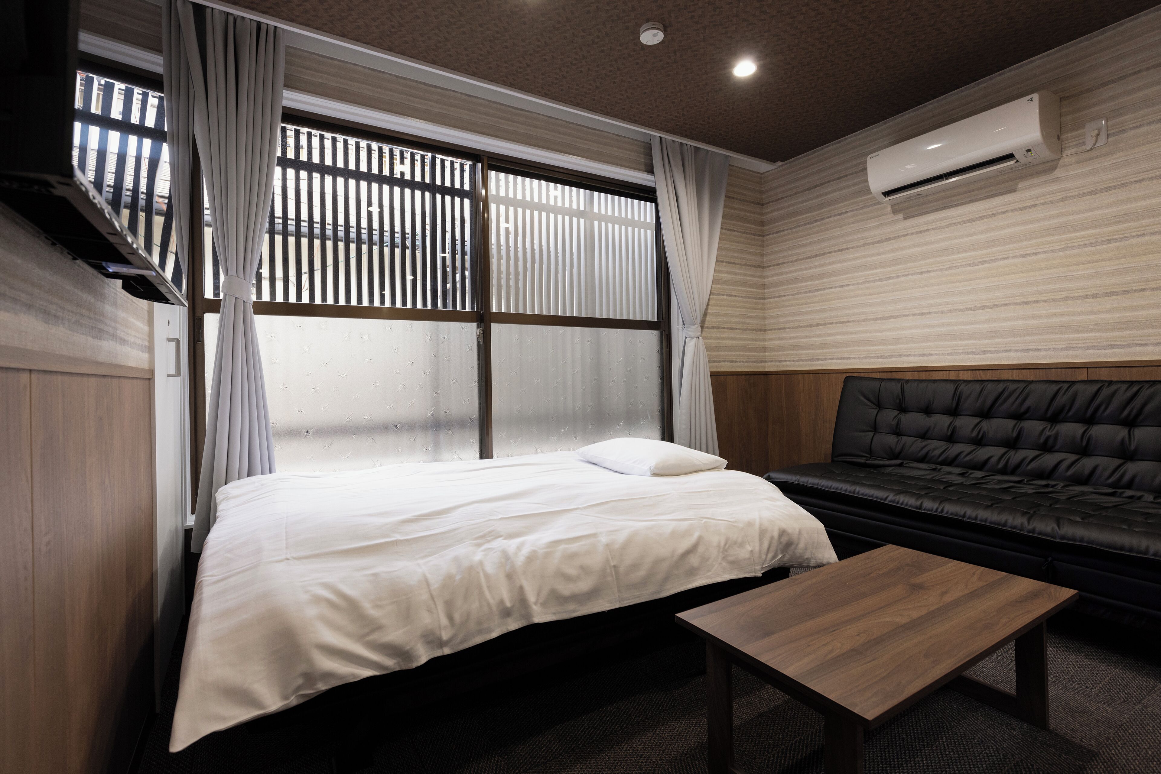 A hotel room with a bed, a sofa, a wooden coffee table, and an air conditioning unit on the wall.