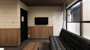 A modern room with a wooden panel wall, a black leather sofa, a flat-screen TV mounted above a wooden cabinet, and a window with blinds. - Whitedragon60 (Osaka)
