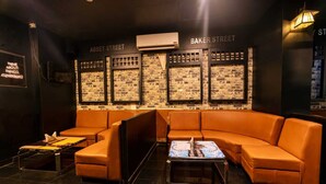Un espacio lounge moderno con un sofá de cuero naranja curvo, una mesita de centro pequeña y una pared con diseño de ladrillo que muestra la señalización 'BAKER STREET'.