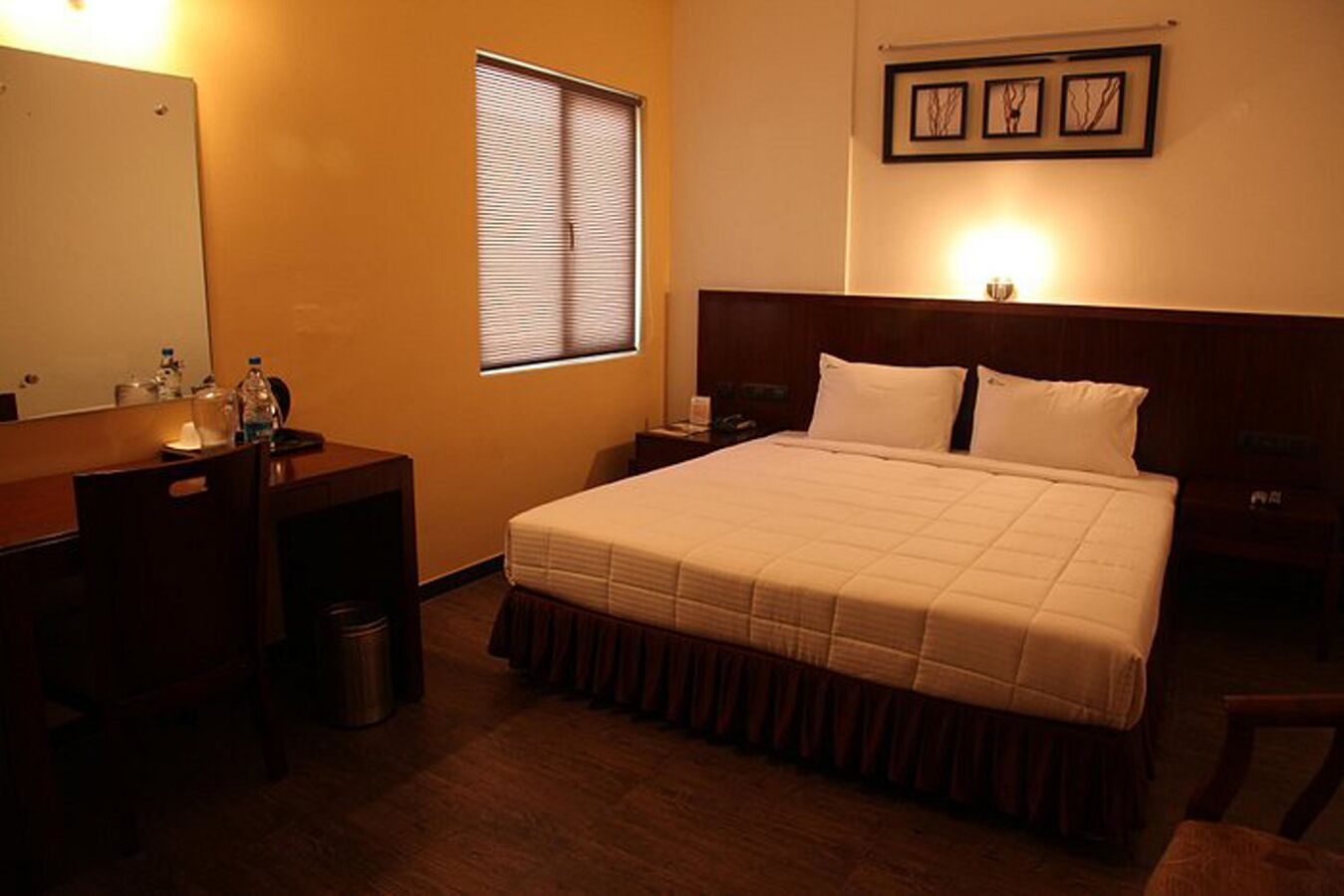 A hotel room with a bed, a desk, a chair, a trash bin, a window with blinds, and framed artwork on the wall.