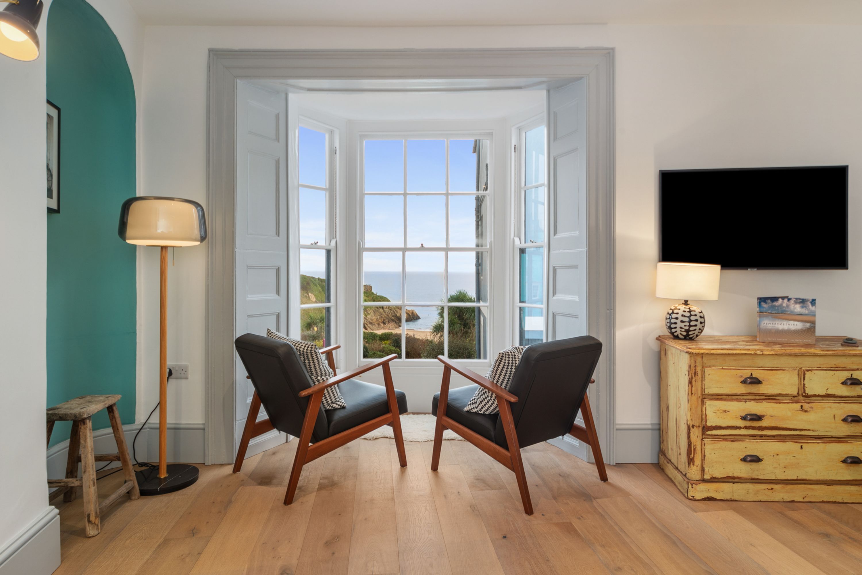 Une pièce avec deux chaises noires, placées face à une grande fenêtre donnant sur la mer. On y trouve une commode en bois avec des tiroirs et une lampe posée sur le dessus.