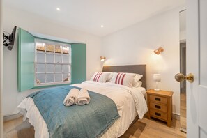 A neatly made bed with a blue and white bedspread, two rolled towels, and a bedside lamp. There is a wooden nightstand with a lamp and a drawer. A window with a green frame is visible, and a door is partially open. - Windermere House - Sea Views Close to Harbour (Tenby)