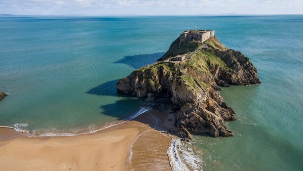A coastal landscape with a sandy beach, a rocky outcrop, and a fortified structure on a cliff. - Windermere House - Sea Views Close to Harbour (Tenby)