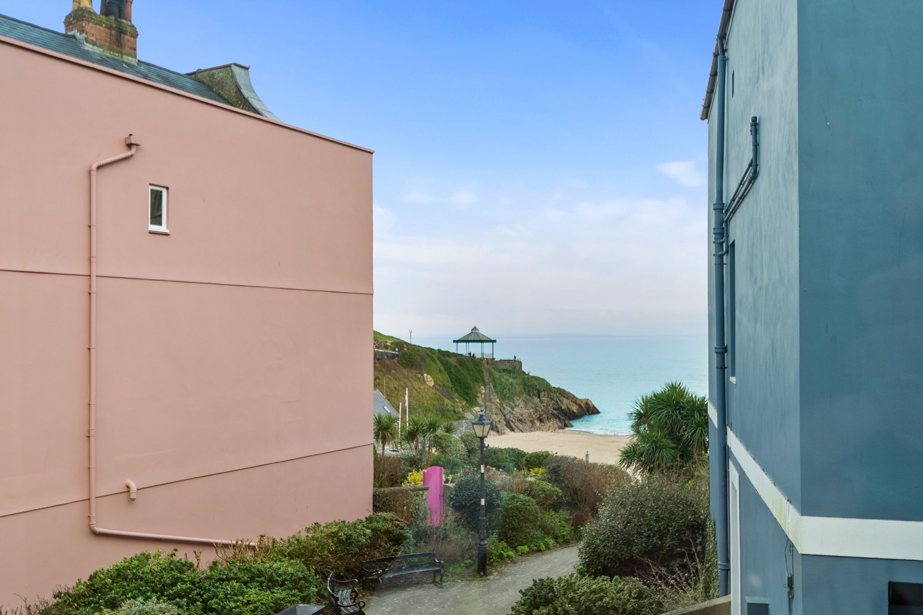 A coastal view with a path leading to a beach, a gazebo, and a building with a pink facade.