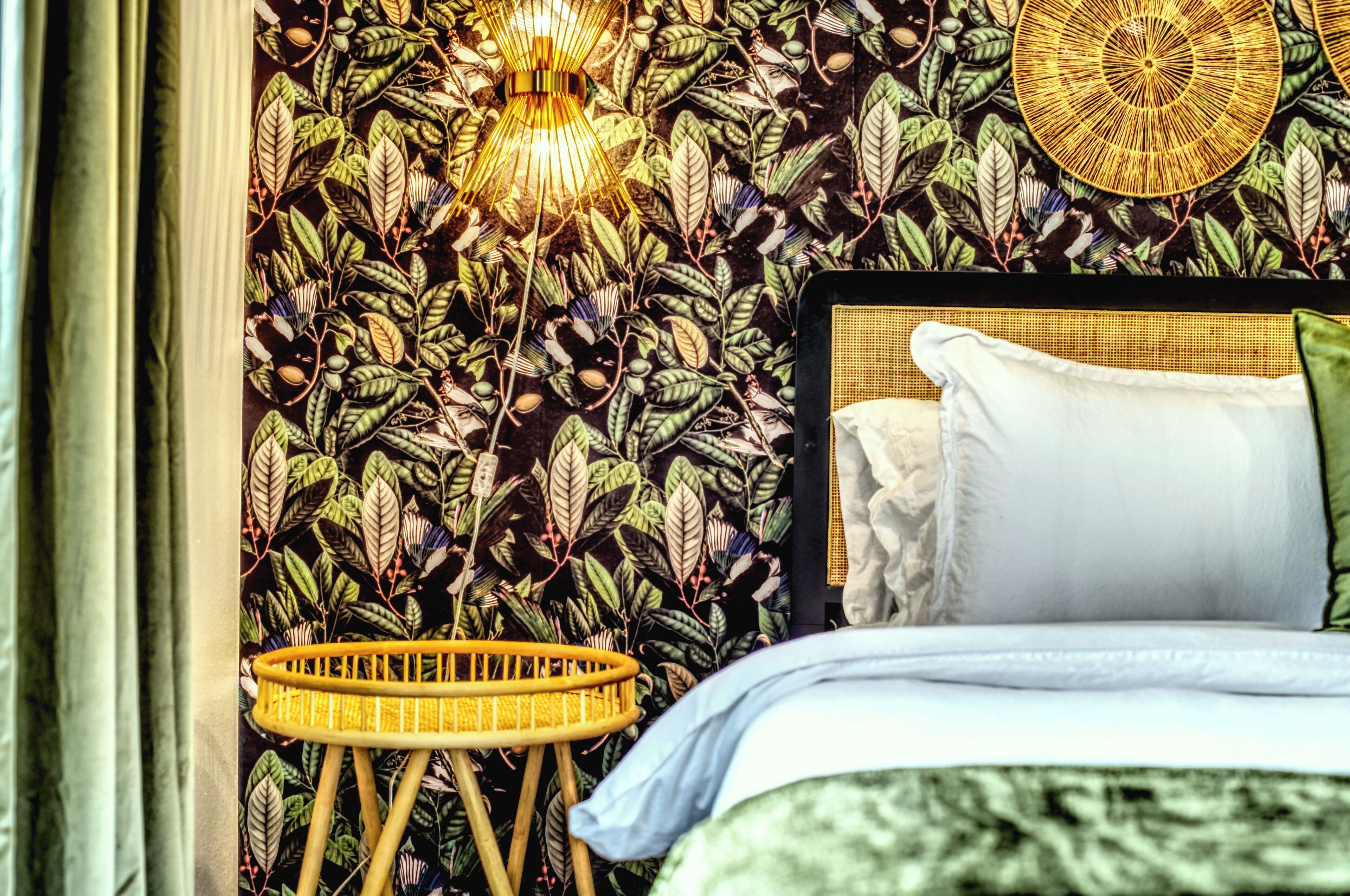 Ein Schlafzimmer mit einem Bett, einer Wand mit Blättermotiv, einem Korbstuhl und einer Pendelleuchte.