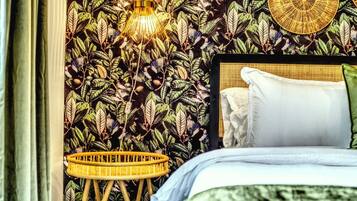 Ein Schlafzimmer mit einem Bett, einer Wand mit Blättermotiv, einem Korbstuhl und einer Pendelleuchte.