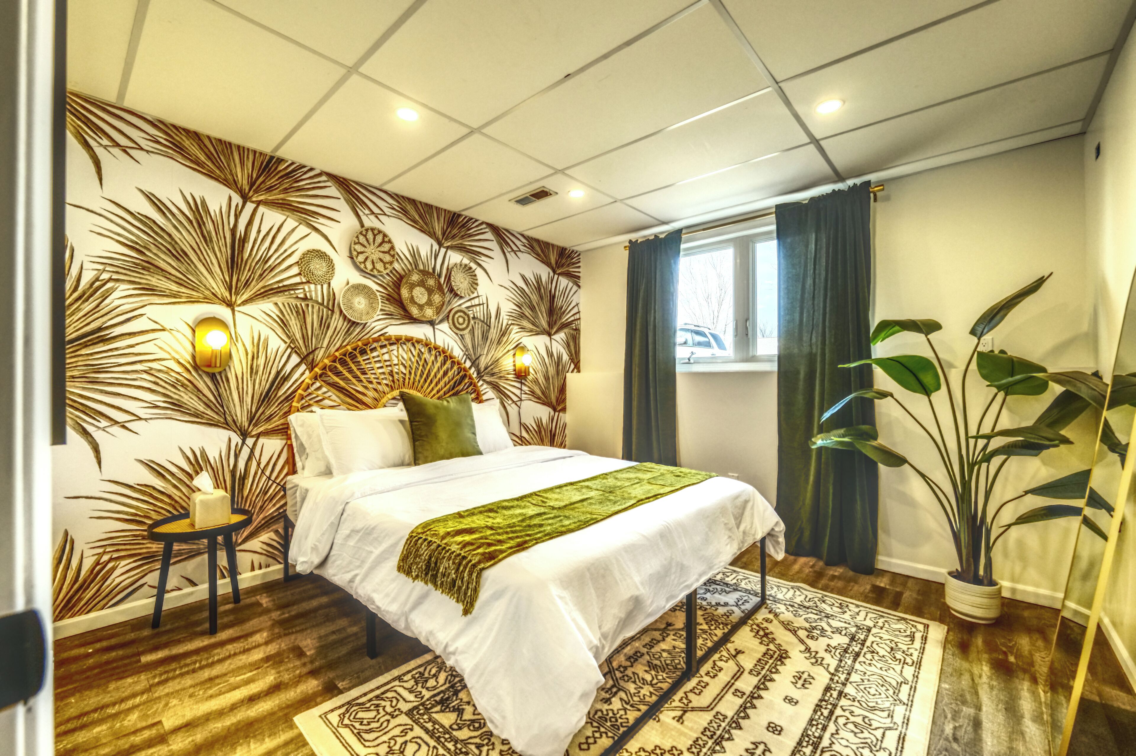 Een hotelkamer met een bed, nachtkastje, een bloempot en een raam met gordijnen.