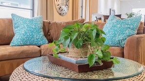 Een woonkamer met een bruine bank, gekleurde kussens, een glazen salontafel en een plant in een pot.