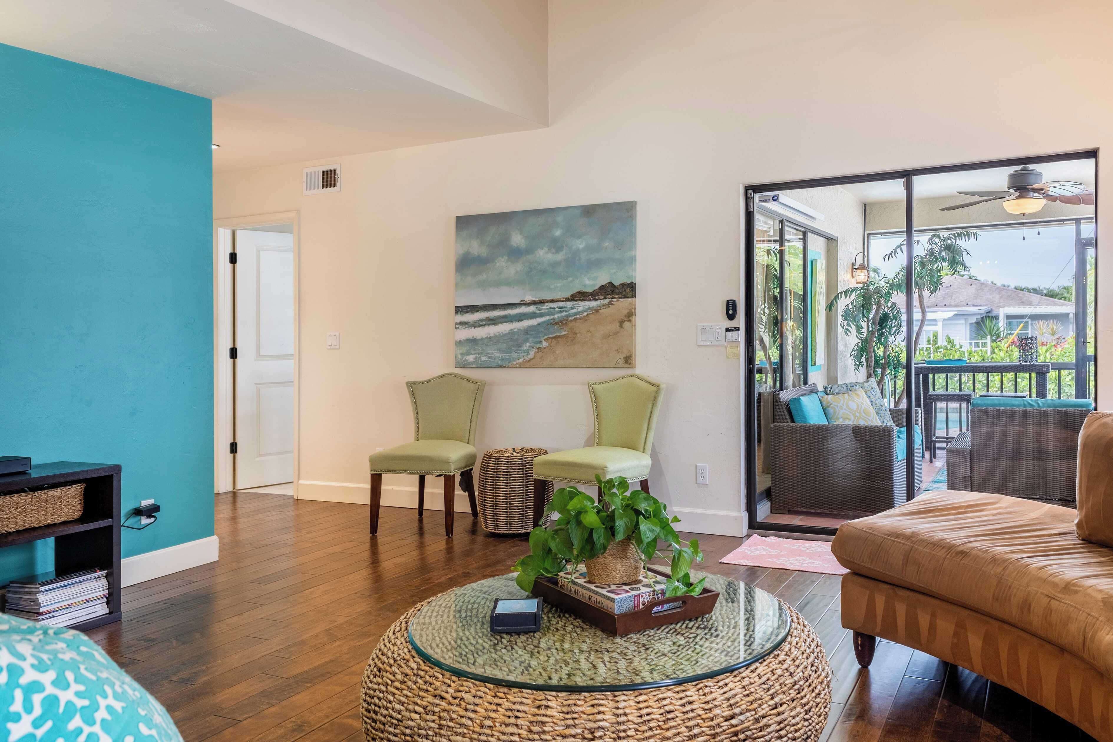 A living room with a large window, a painting of a beach, and a view of a house.