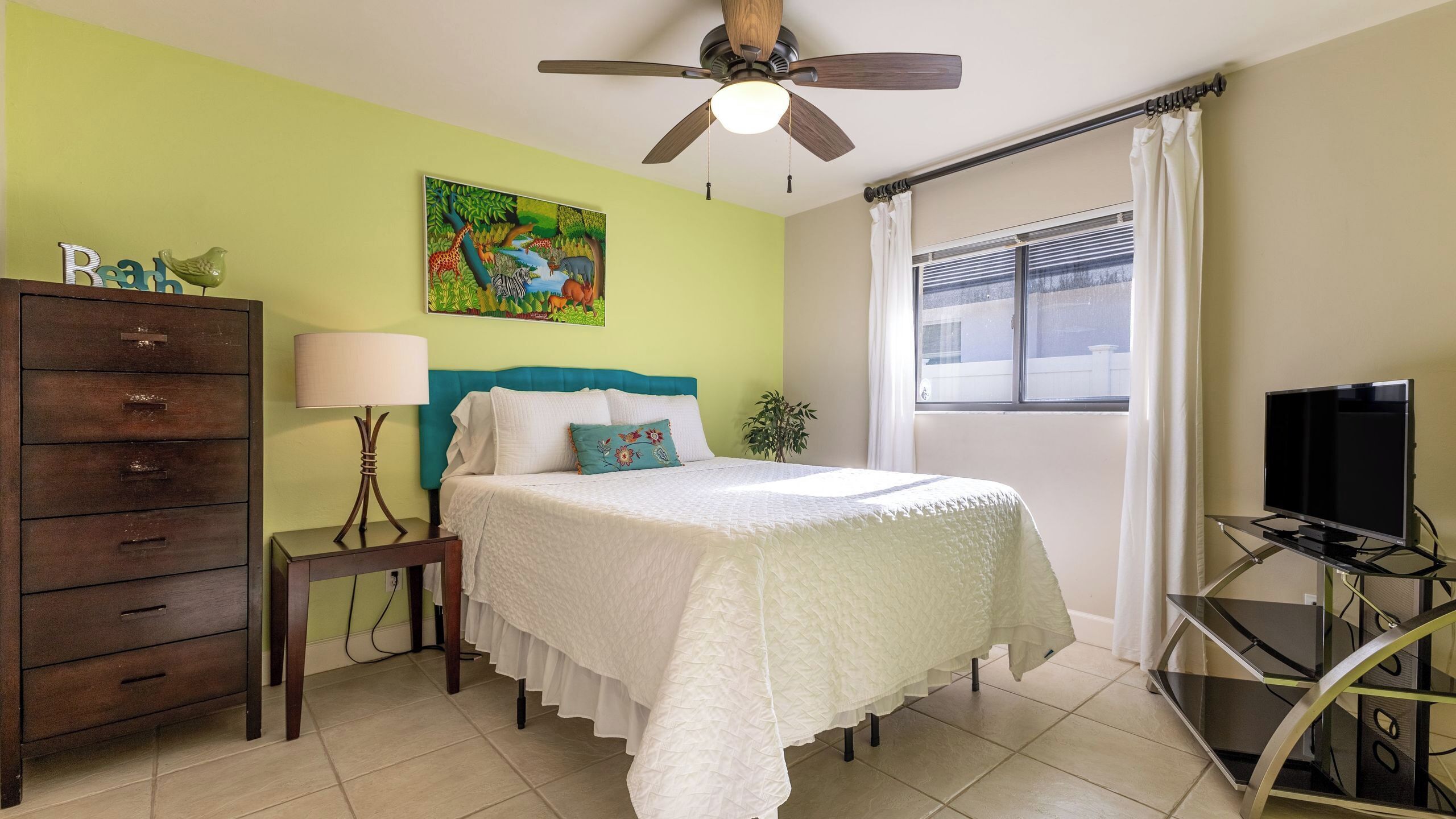 A bedroom with a bed, a dresser, a lamp, a painting, a window, and a TV on a stand.