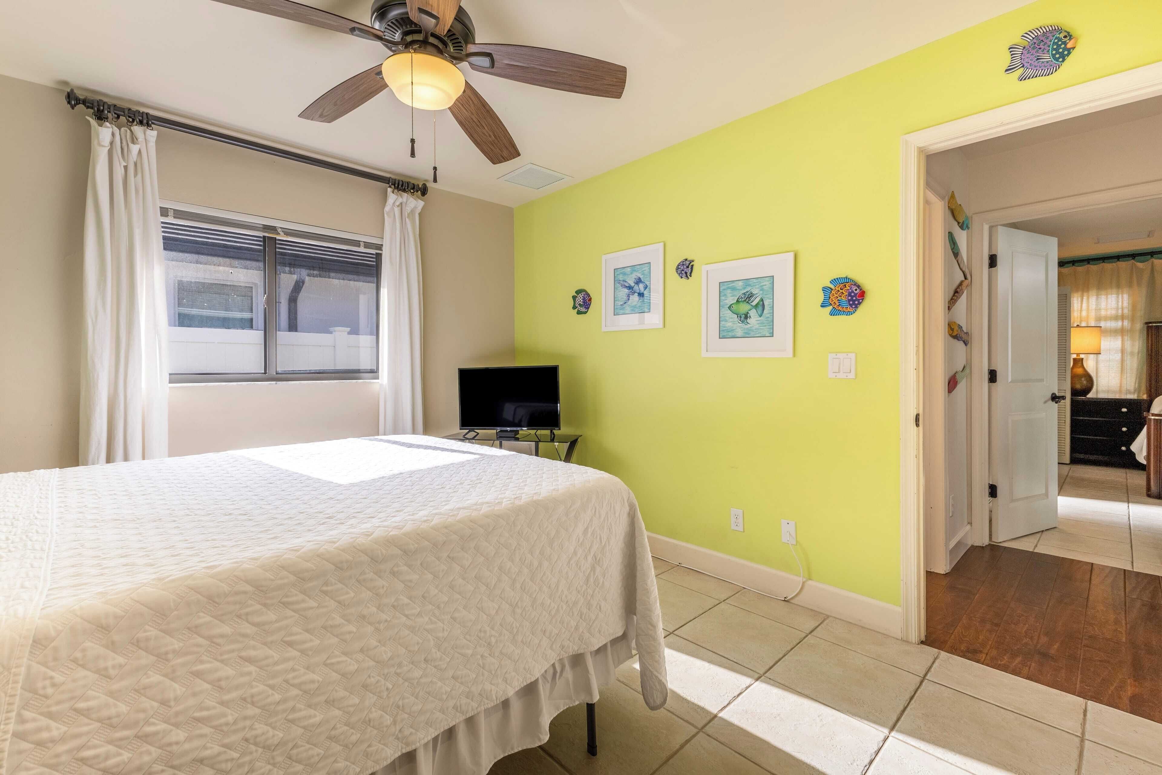 A bedroom with a bed, a ceiling fan, a television, and a door leading to another room.